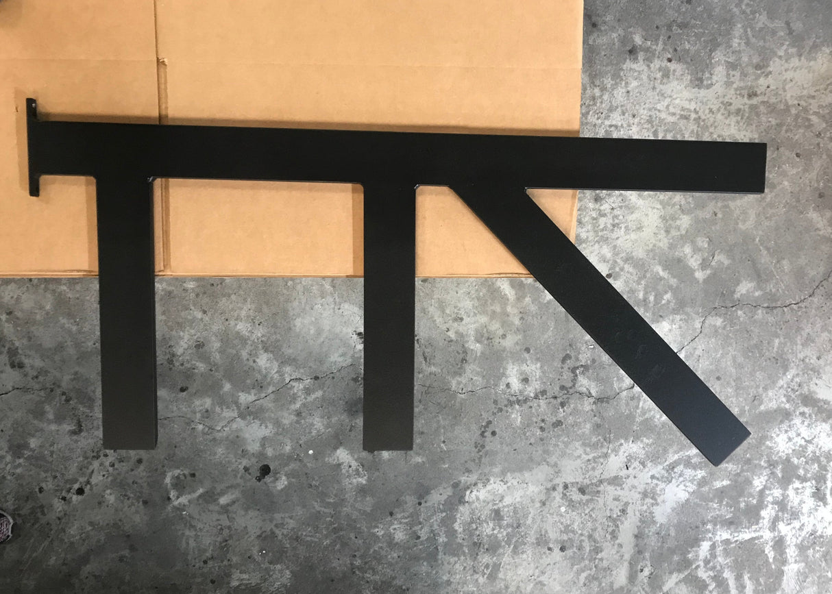 A Long Black Custom Support Bracket with two arms coming down vertically and one arm with a 45-degree angle from the top, sitting on a cardboard box on a cement floor.