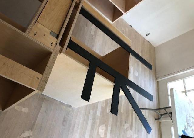 Top-down angle of Black Custom Support Brackets sitting on an unfinished wooden countertop with a white work table in the background.